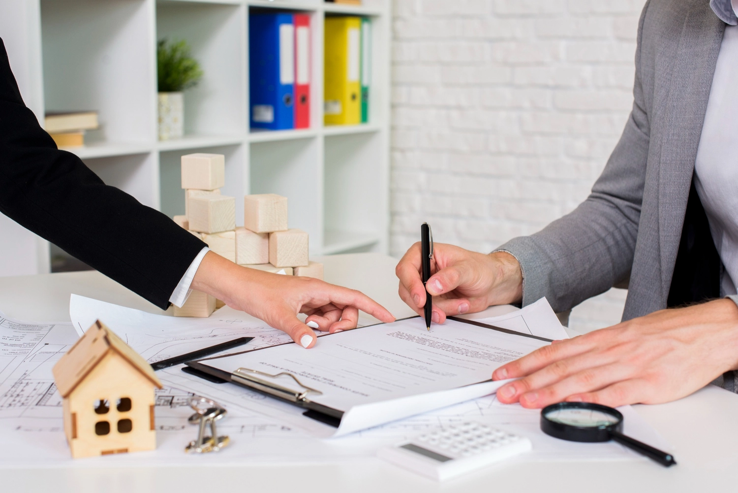 Roland Renner GmbH - Immobilienverwaltung und Gebäudedienstleistungen - Eine Person in einem grauen Blazer unterschreibt ein Dokument auf einem Schreibtisch, während eine andere Person in einem schwarzen Blazer auf eine Zeile auf dem Papier zeigt.
