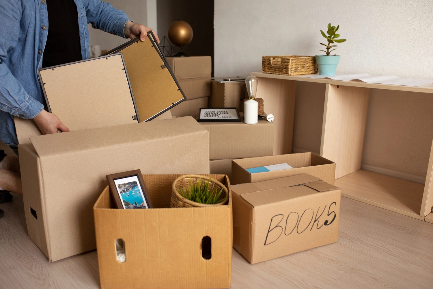 Roland Renner GmbH - Immobilienverwaltung und Gebäudedienstleistungen - Eine Person packt in einem Zimmer mit einem hellen Holzregal gerahmte Bilder und Haushaltsgegenstände in mehrere Kartons.