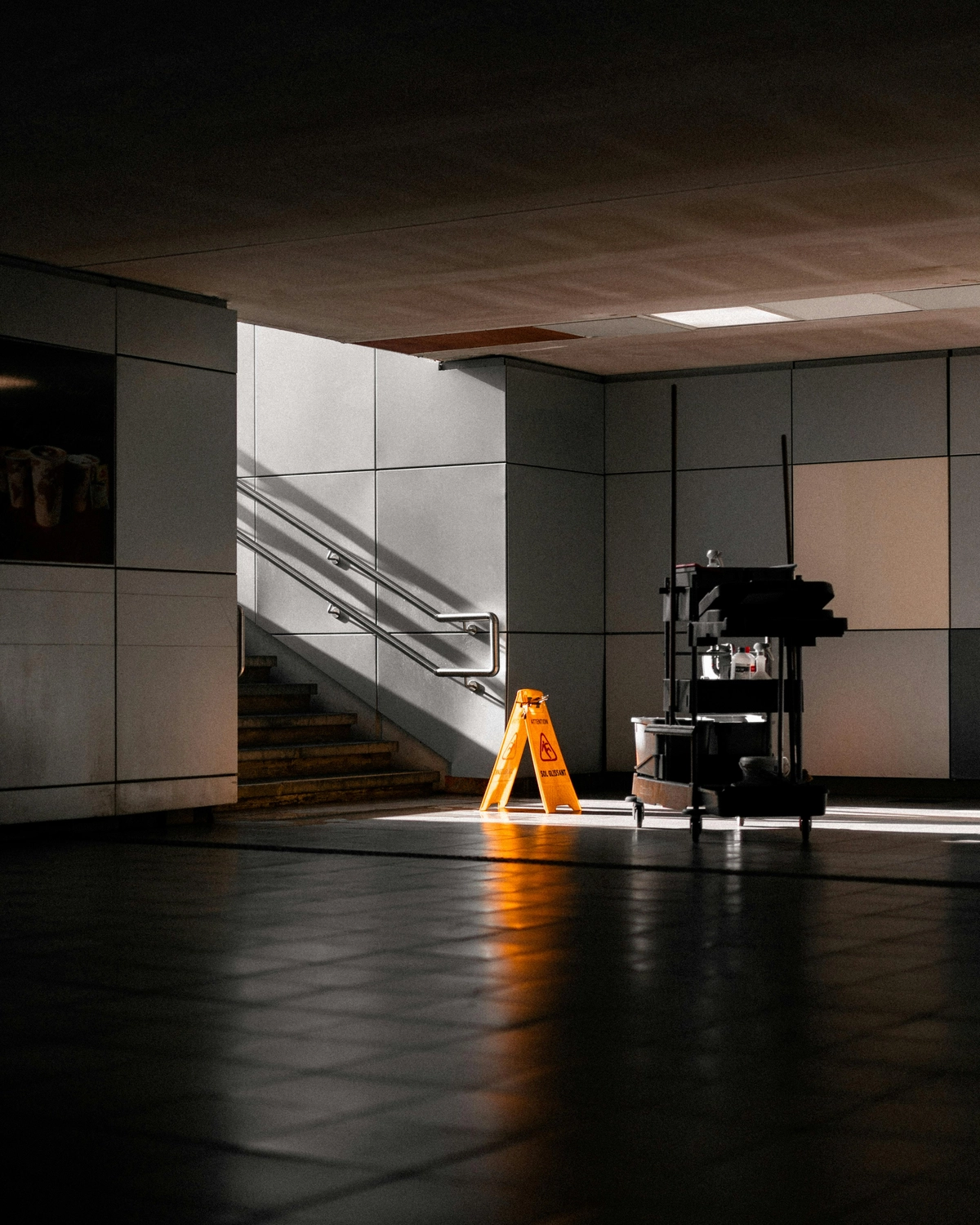 Roland Renner GmbH - Immobilienverwaltung und Gebäudedienstleistungen - In einem gefliesten Flur in der Nähe einer Treppe stehen ein Reinigungswagen und ein gelbes Warnschild „Vorsicht, nasser Boden“.